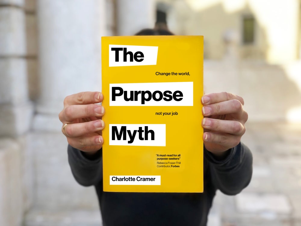 Close-up of hands holding a book to camera. The book has a yellow cover and is titled 'The Purpose Myth' 