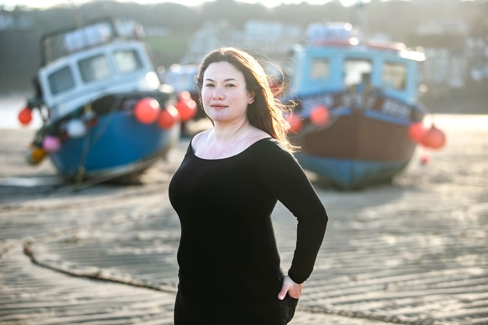 A woman stands in front of 2 boats on the beach.