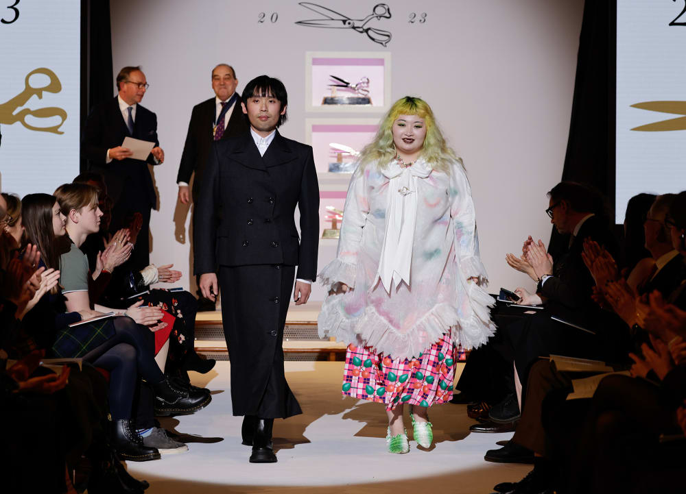 Image shows 2 people walking down a fashion runway with a spotlight on them and audience on either side. The person on the left is wearing a black suit top and long black skirt. The person on the right is wearing a pink skirt, with yellow hair and floaty off-white top. The audience is clapping.
