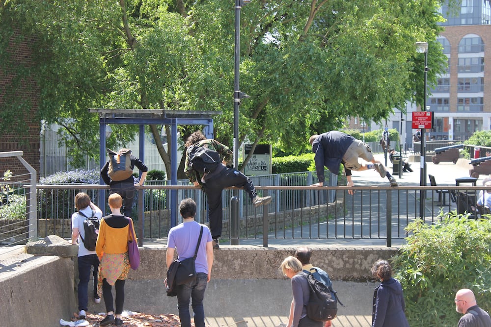 Three people jumping over a metal fence in an urban environment. A line of more people are following them.