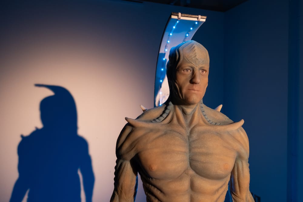 Image shows a life-size model of a blue, shimmery, alien with spikes on its shoulders and a tall apparatus above its head