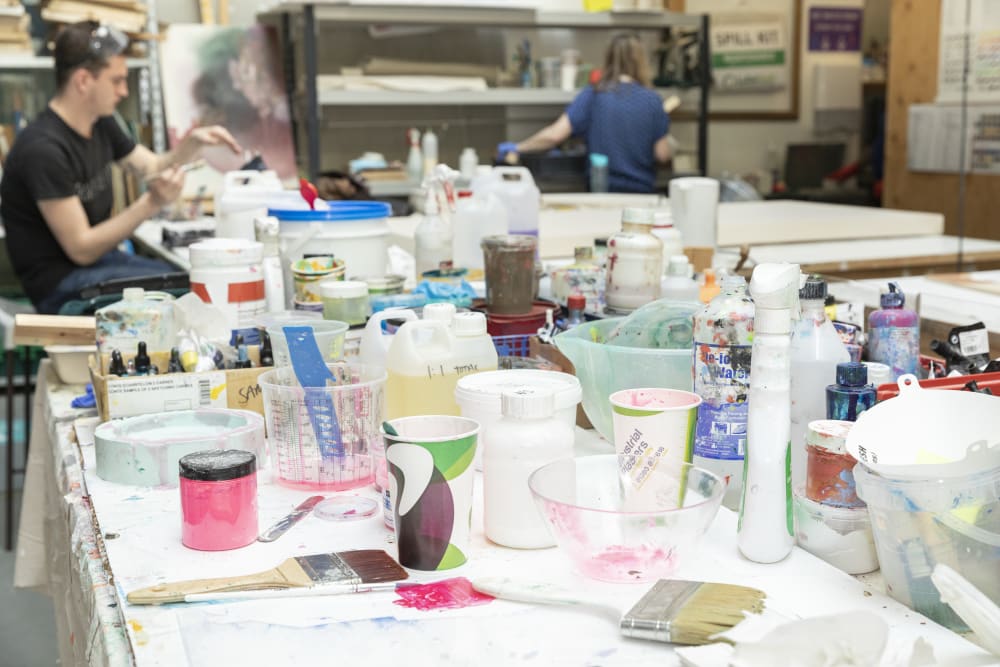 Close-up on a table in the Paint workshop, with a variety of paints, materials and utensils. Students are visible working at the edges