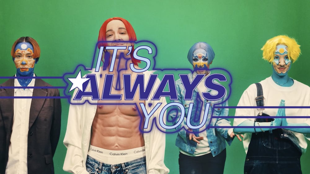 A still from 'It's Always You'. There are four people against a green background wearing theatrical makeup and with brightly coloured hair.