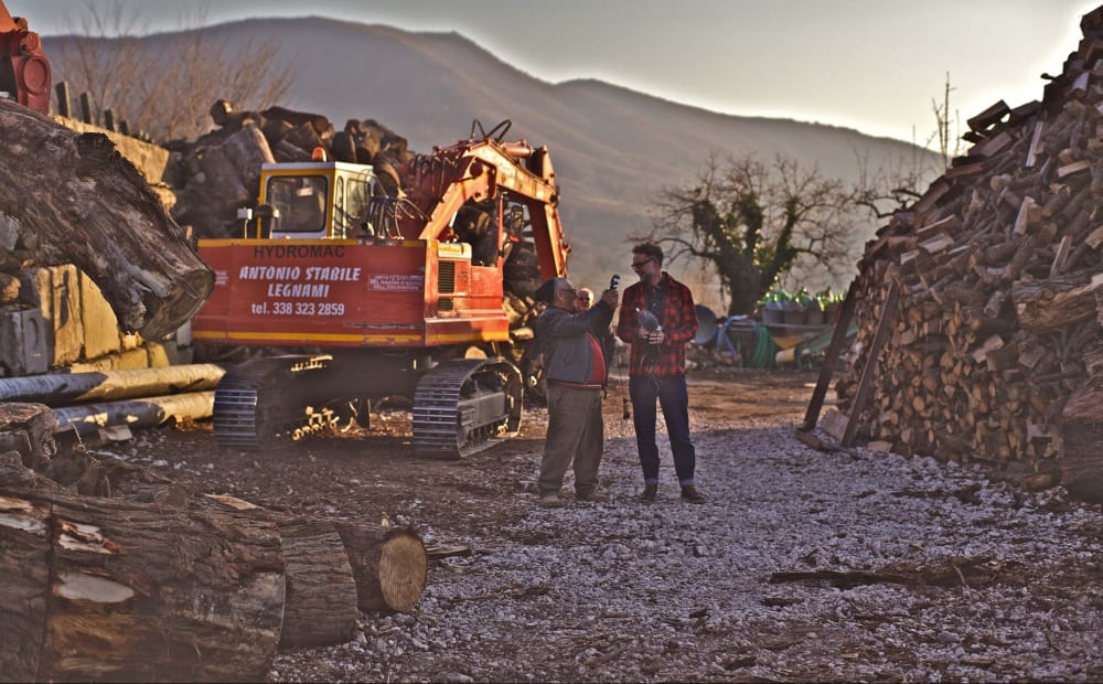 Two people standing in a timber yard next to a large digger. One of them is holding a microphone. In the background are mountains.