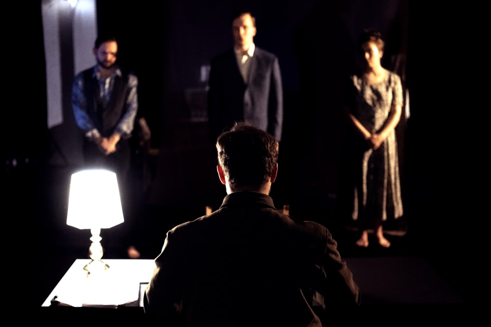 A man is sitting in the forefront with his back to his. He's wearing a suit and lit from a lamp on his lefthand side. He's looking at 3 blurry figures stood further back. 