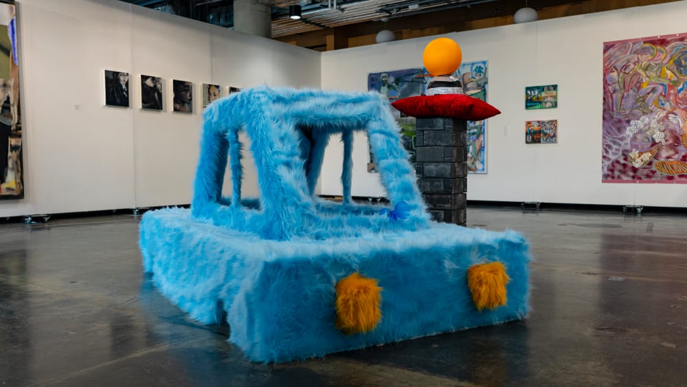 Blue, fuzzy car displayed at an exhibition surrounded by other work hung up on white walls.