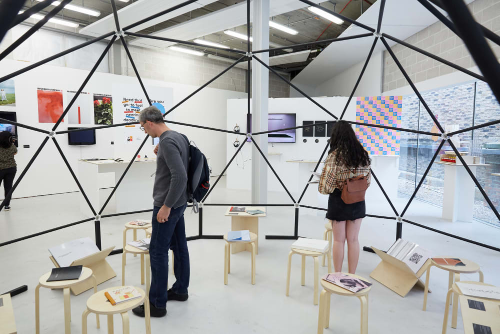 Two people look at work resting on stools in a specially-created reading area at the BA Graphic Design Communcation exhibition at the Chelsea undergraduate Summer Show 2019.