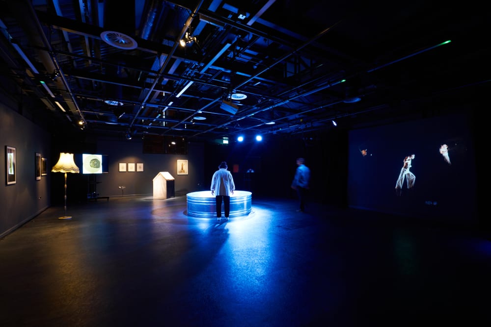 A person stands in the middle of a large studio which is very dimly lit. In the centre, where they are standing, a blue light emanates from a circular piece of art.