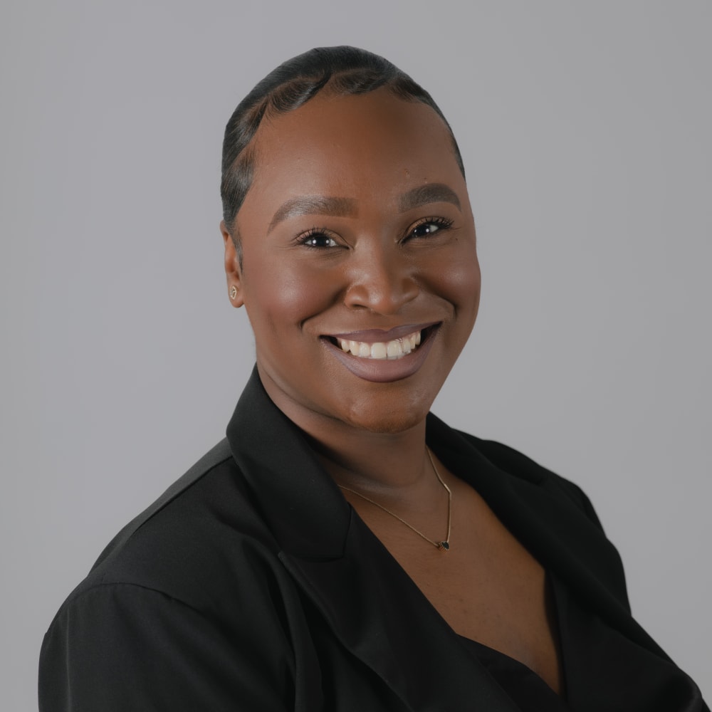 Headshot of a person stood with their body angled towards the camera. They are wearing a blazer, smiling and lit by studio lighting with a plain grey backdrop.