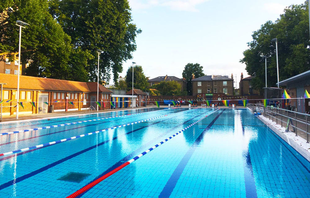 An outdoor swimming pool with lane dividers, surrounded by buildings, trees, and fencing, photographed on a clear day.