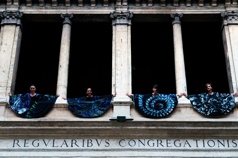 Figures wear blue while standing in a Greek-style building.
