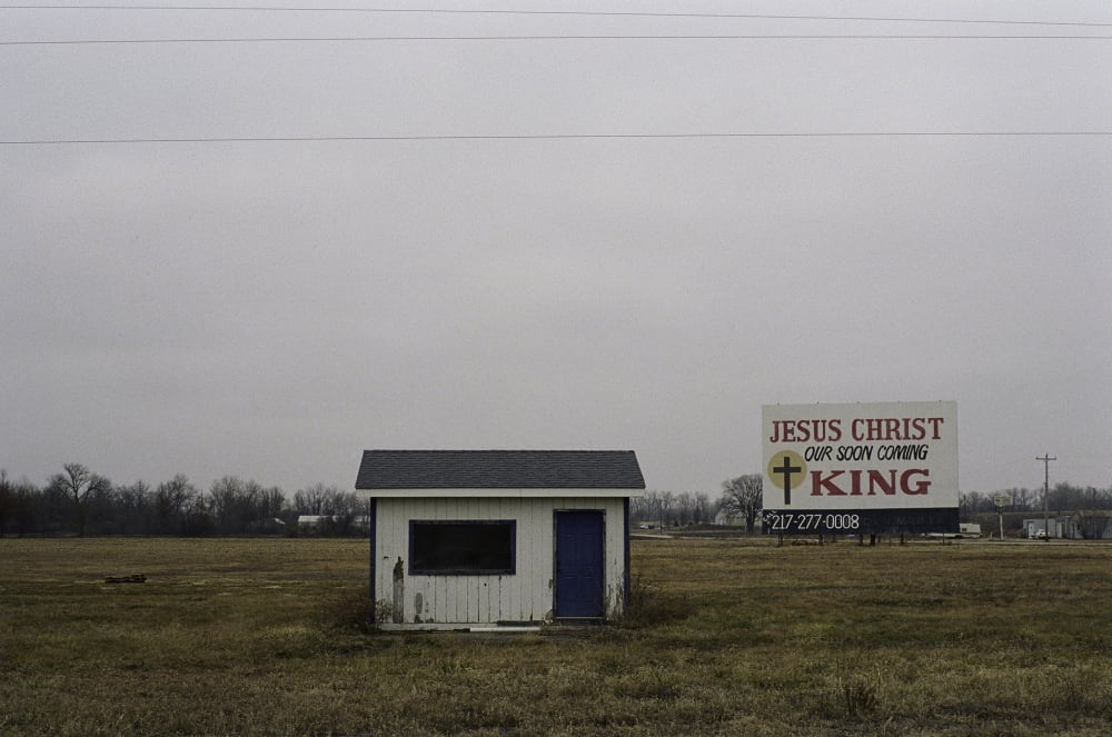 Photograph of a small building by a road, sign nearby reads Jesus is King