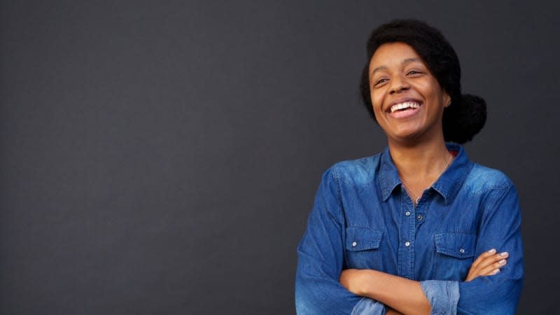 A woman in a denim jacket with her arms folded in front of a grey background - she is smiling. 