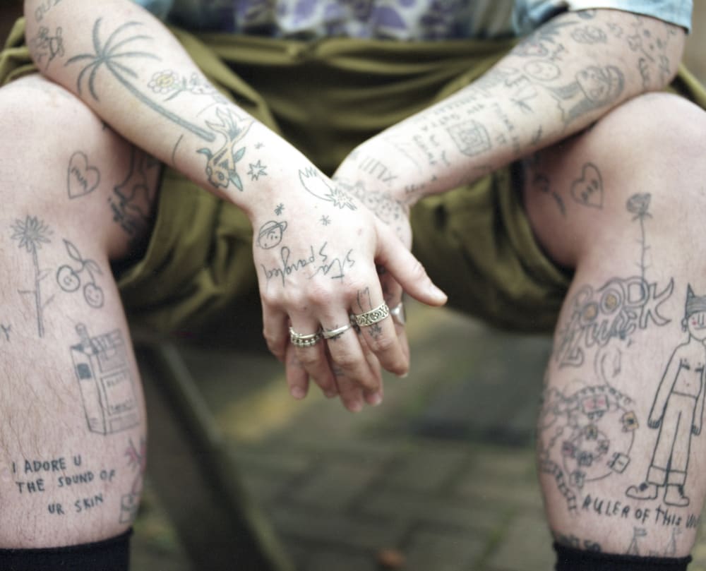 A person wearing shorts rests their hands over their thighs. Their legs and arms are covered in tattoos