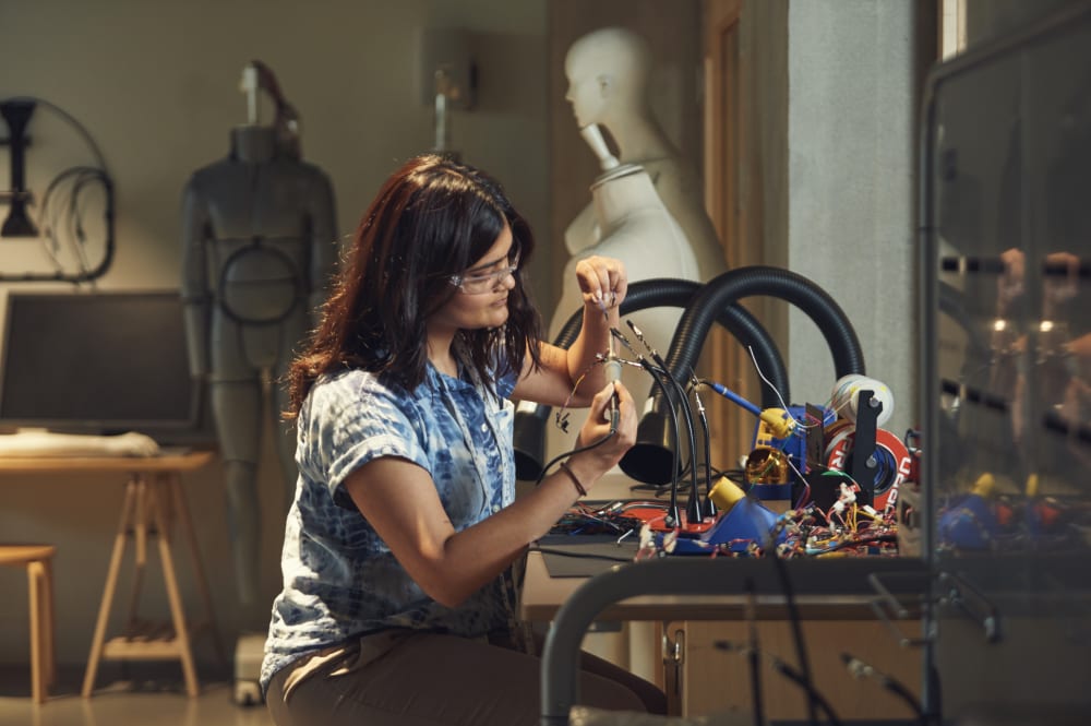 Student wearing safety glasses working on an electronic project at a cluttered desk with wires and tools, in a workshop with mannequins and equipment in the background