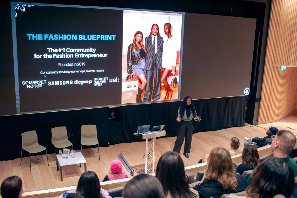 Wide shot of Giovanna presenting with guests seated in front of her. On the screen behind her it says: THE FASHION BLUEPRINT, the #1 community for the fashion entrepreneur. Founded in 2019, consultancy services, workshops events + more.' Underneath, are the logos for: Somerset House, Samsung, Depop, Graduate Fashion Week and UAL.
