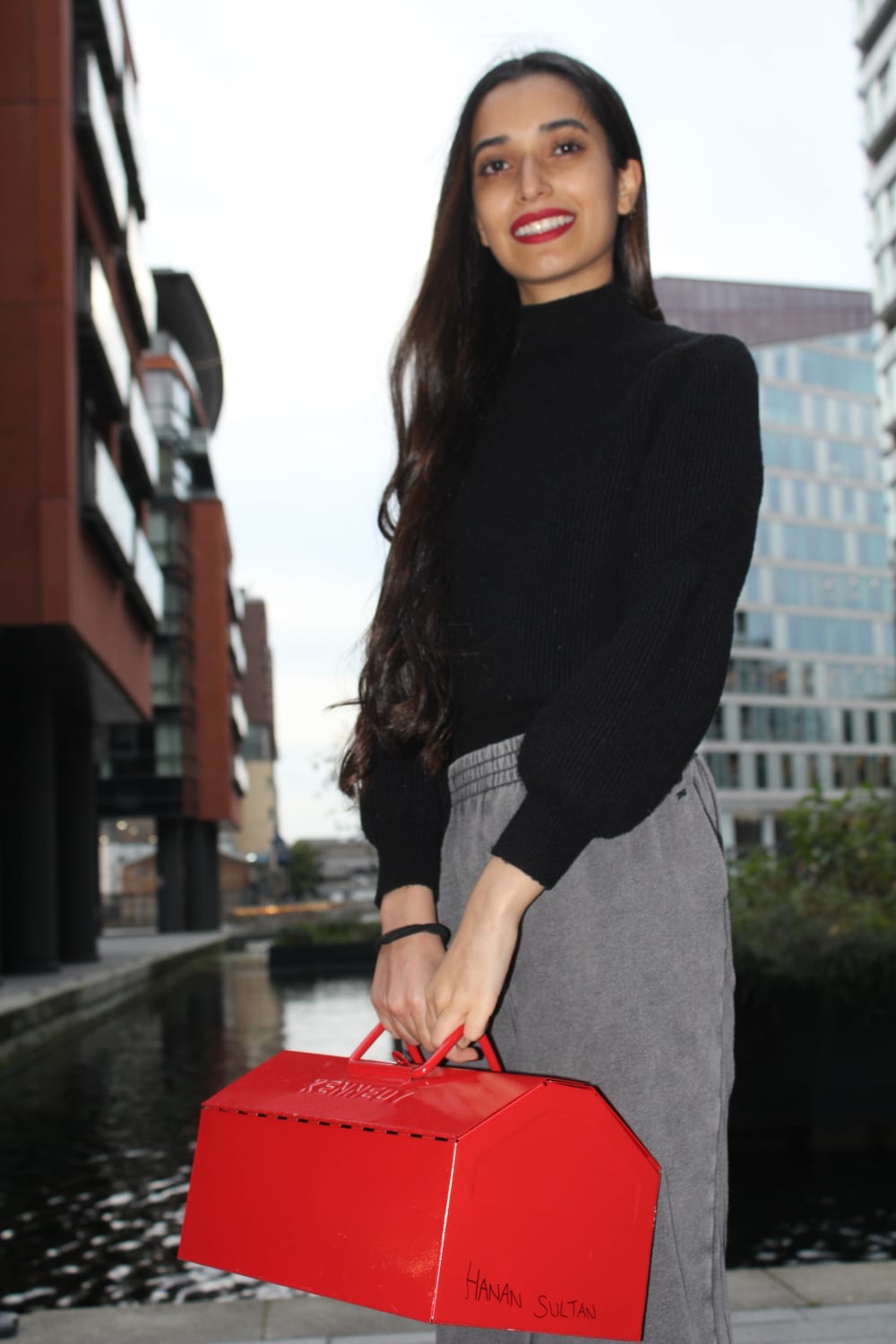 Hanan standing and smiling into the camera. She's wearing a black polo neck, grey trousers and is holding a bright red tool box