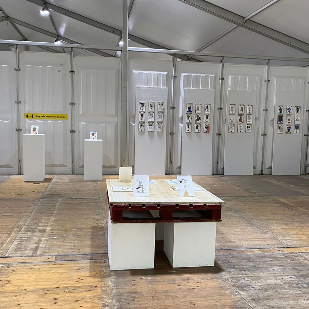 Image is of inside the Chelsea tent, centre there is a table with art works positioned upright and flat, in the background there is a wall, on the right of the wall there are 3 panels each panel has 9 small portraits on. On the left in front of the wall are two white podiums each with a portrait positioned upright on top.