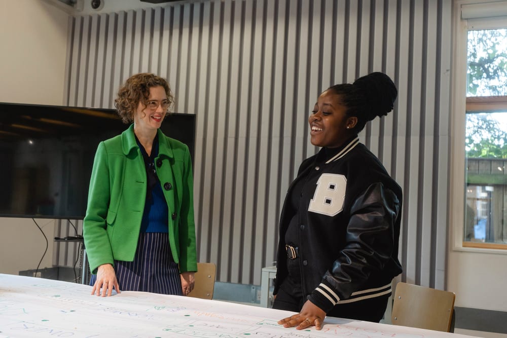 Image shows a young person on the right hand side speaking with a woman on the left hand side. The woman is wearing a green jacket and wears glasses. The young person is wearing a black jacket with the letter B detailed.