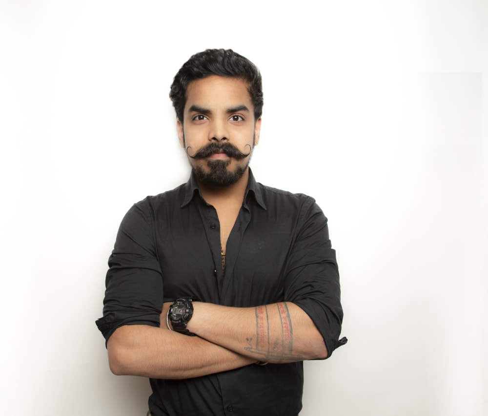 Shubhangam Singh, wearing a black shirt with his arms folded, staring into the camera. He has a handlebar moustache and a tattoo around his left forearm.