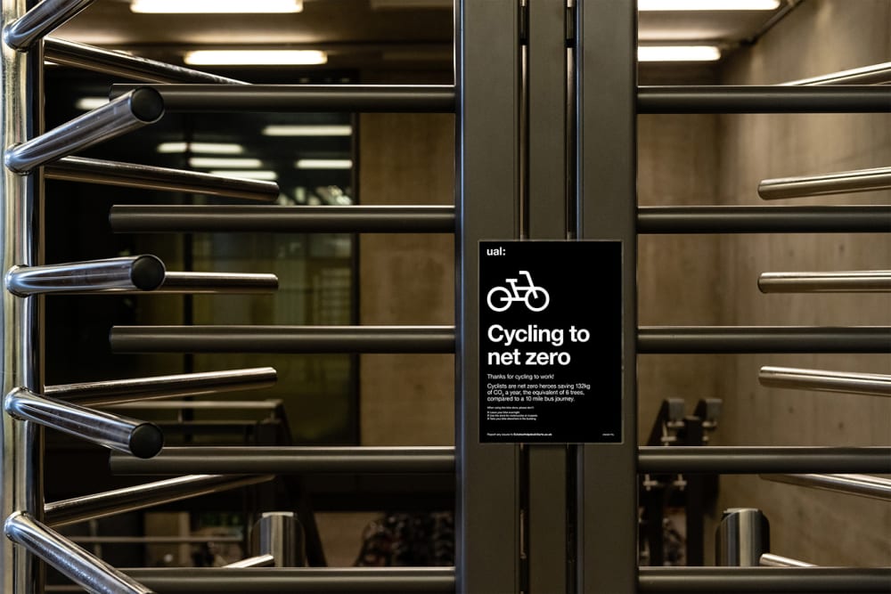 Close up photograph of the metal turnstiles entrance to bike storage. There is a black background poster stuck on the metal post with a white bike icon at the top which reads 'cycling to net zero'.