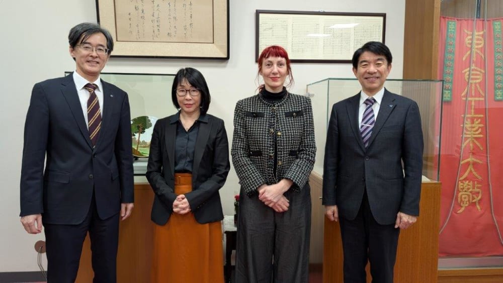 Four people posing in front of an exhibition with framed posters and objects behind rectangular glass.