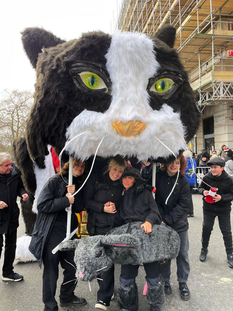 Image shows 4 people in the foreground holding up a puppet of a large black and white cat head, with two people in the background on either side of the cat.