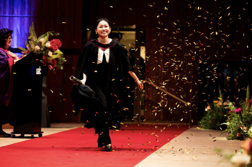 A student walking down a red carpet on stage surrounded by blue and yellow confetti in the air. Their face and body are lit by spotlights and they are smiling into the top right corner of the image. There are flowers in the background decorating the stage,