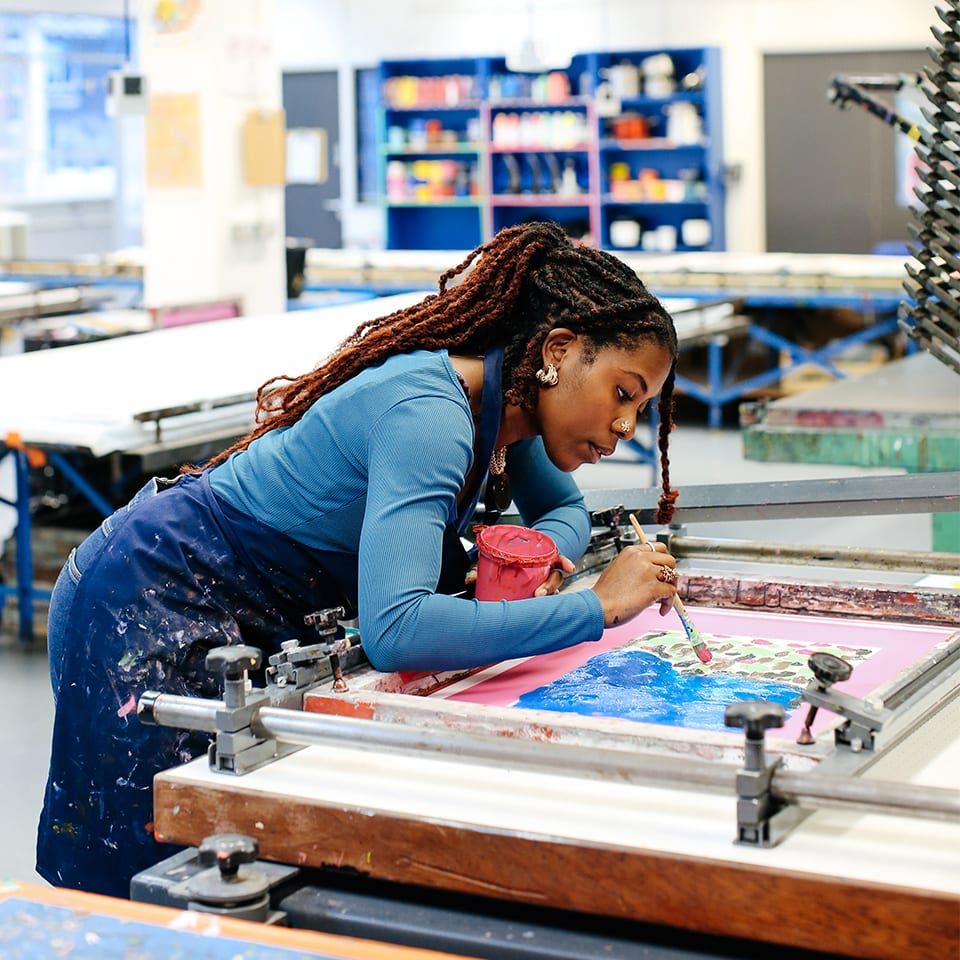 A student Davinia Clarke leans over a printing workshop table, painting onto a silk screen.