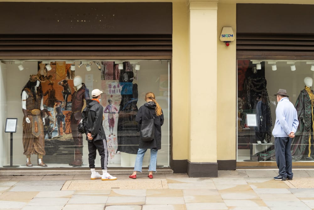 three people have stopped in the street and are standing with their backs to the camera, looking in detail at student costume work in the windows of two large shopfronts.