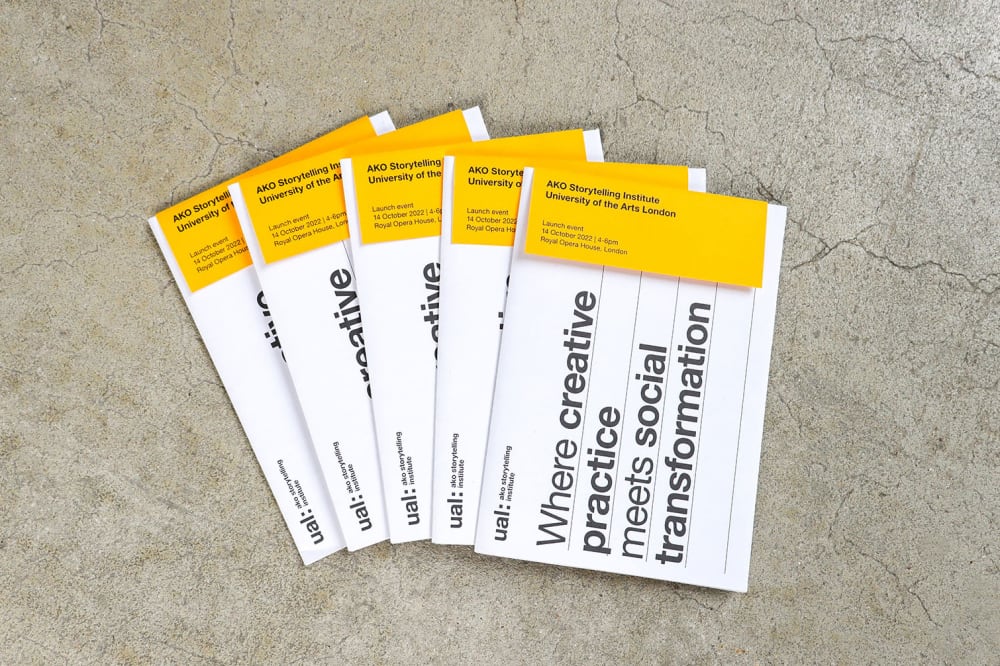 5 printed booklets laid out in a fan on a concrete floor. The booklets read 'UAL AKO Storytelling Institute, where creative practice meets social transformation' and they have a yellow insert hanging over the top of the front page.