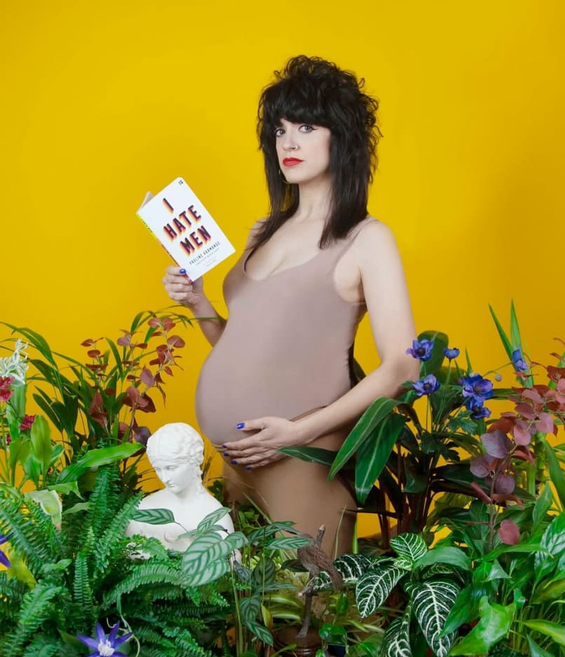 A portrait of a pregnant Sarah one hand on belly the other holding a book titled 'I hate men'. Sarah is standing amongst greenery and a white bust statue against a yellow wall.