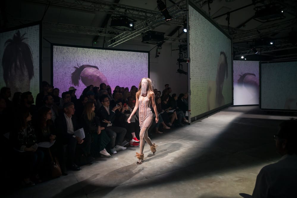 Model walks down a catwalk. Several large screens surround the catwalk displaying faceless heads.