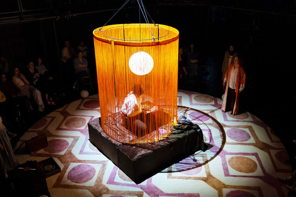 Image shows a large circular orange fringed lampshade with a white light inside. A person can be seen sitting in the middle of the orange fringe, on top of a square brown seat. A 1970s style rug sits across the floor and another person looks on from the right hand side.