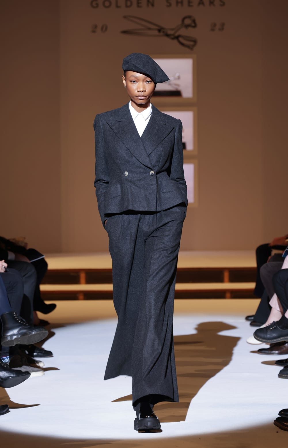 Image shows a person walking down a fashion runway wearing a black suit and black hat with a white top. The person has their hands in their pockets.