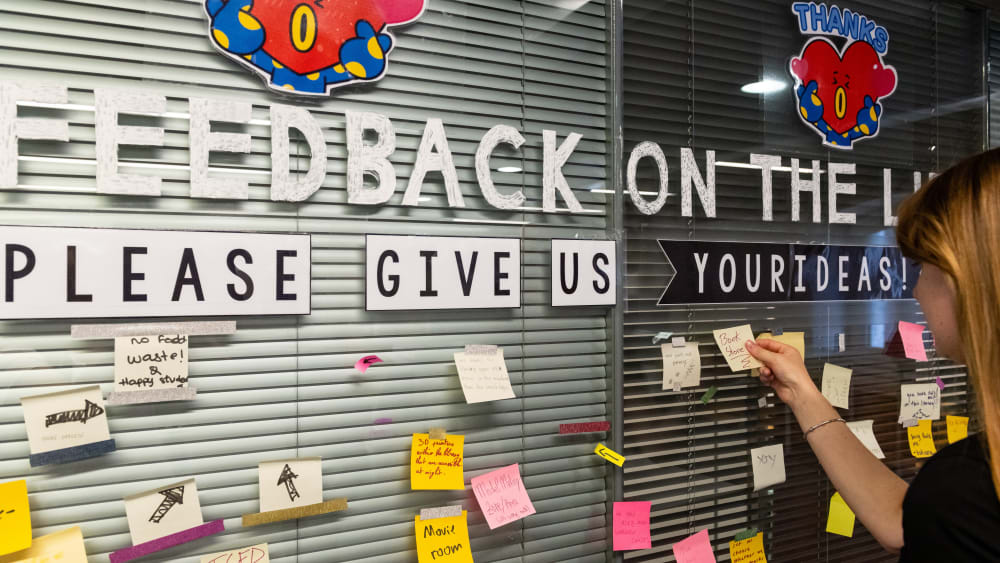 Student placing post it note on glass wall containing more post it notes with feedback suggestions