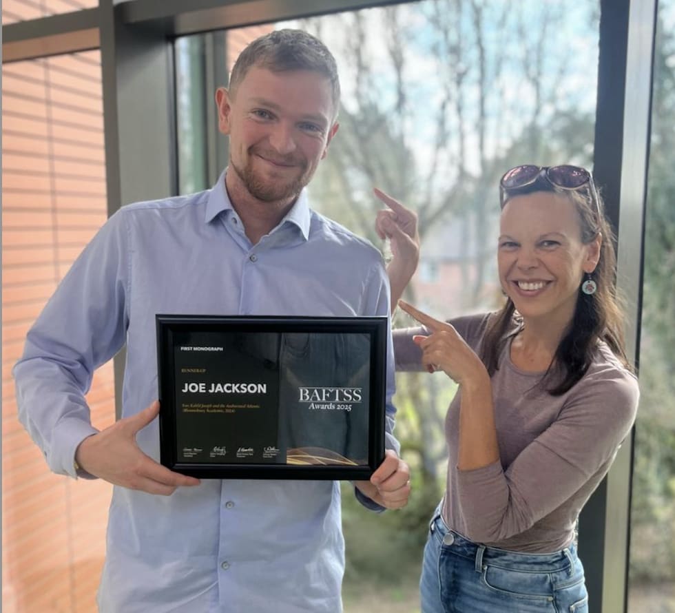 Joe Jackson, a white man with facial hair in a blue shirt poses with his BAFTSS award next to a woman smiling and pointing in his direction