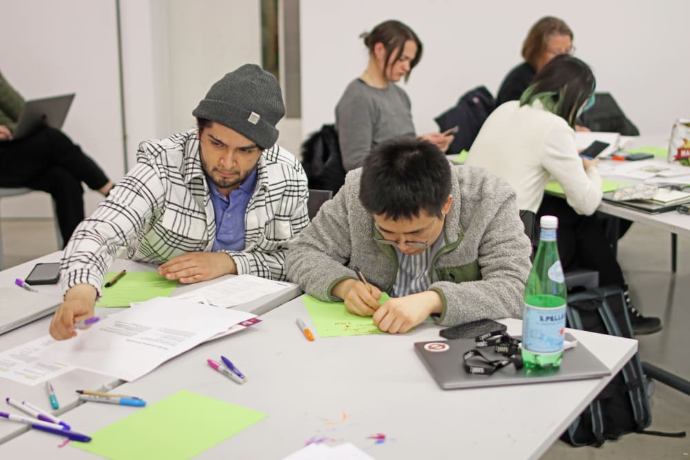 Two students sit at a table and work on their recipe ideas. They are writing and drawing on pieces of paper.