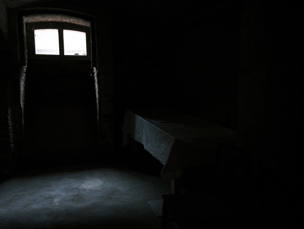 A dark, cold looking room with a stone floor. There is a small window high up on the wall with some light streaming through.