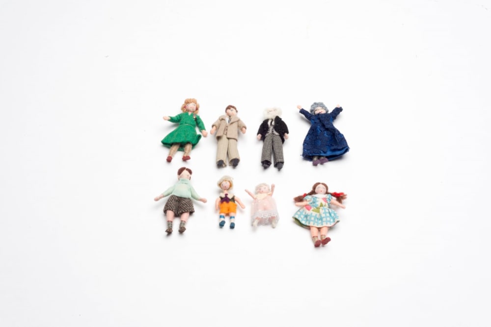 8 miniature cloth dolls are laying down and lined up in 2 rows of 4. Top row, from left to right: a doll in a green knee length dress with blonde hair, a doll in a beige suit and short brown hair, a doll in chequered trousers, black jacket and white shirt with white hair, a doll with a long dark blue dress with grey hair and glasses. Bottom row, from left to right, these dolls are smaller as they are children: a doll with brown shorts, knee length socks, green jumper and brown hair, a doll with orange shorts, brown t shirt and blonde hair, a baby doll in a pink dress, a doll in a cheque blue and white dress with brown hair in pig tails.