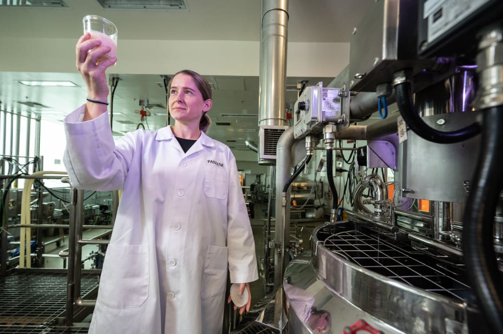 Scientist holding up a clear pot with liquid in. She's standing in a lab, and on her white lab coat it says 'PANTENE'. 