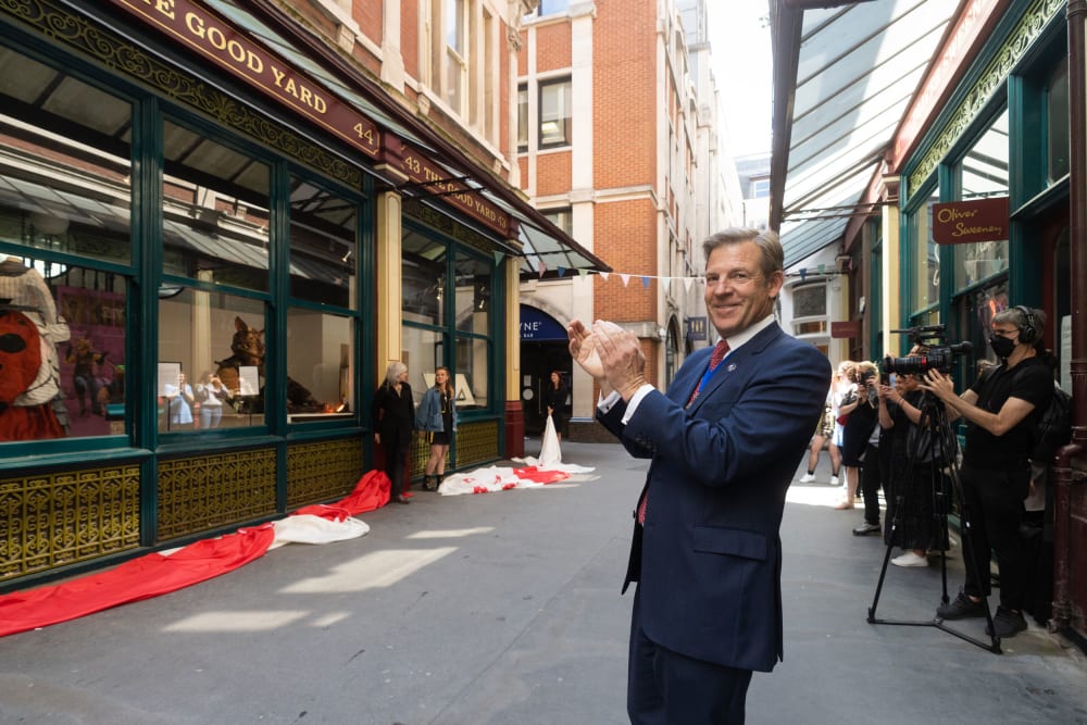 A man in a suit stands, clapping, in front of a row of cameras as students present their work in shop windows to the left of the frame.
