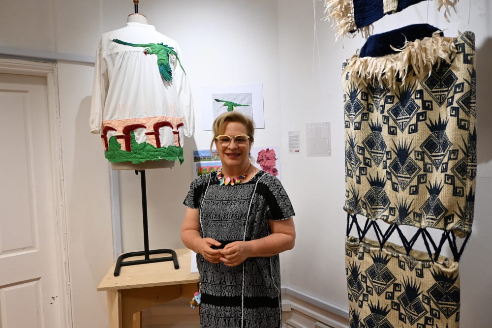 Image shows a woman in a patterned dress with patterned earrings and glasses smiling at the camera in a gallery setting surrounded by textile design work.