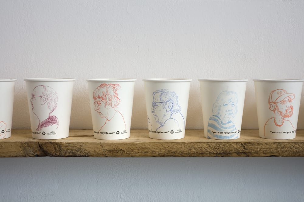 Five paper cups lined up on a wooden shelf. Each as a detailed line drawing of a person in either orange, red or blue ink. 