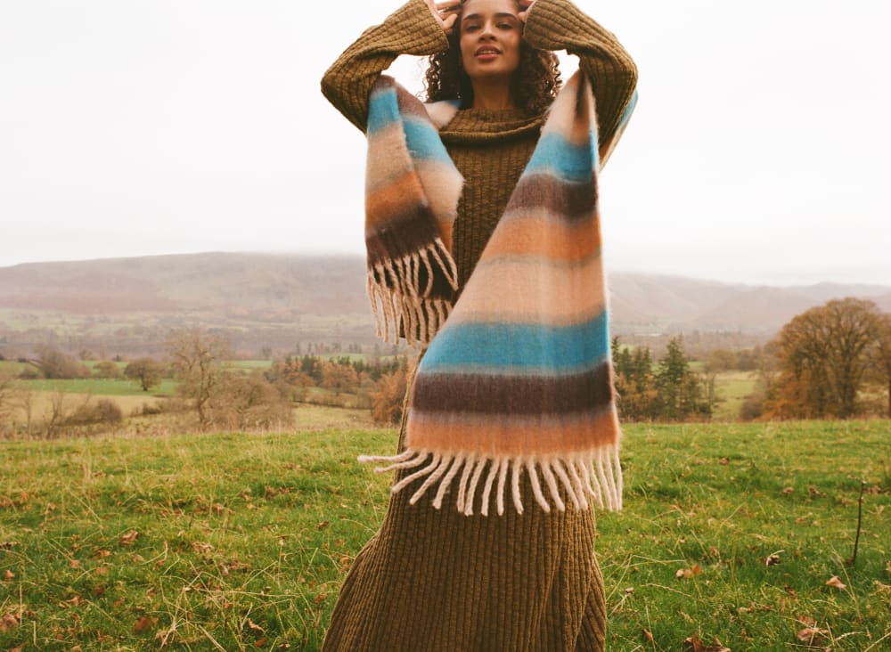 Close up shot of woman standing in a field wearing a khaki knitted dress and a neck scarf