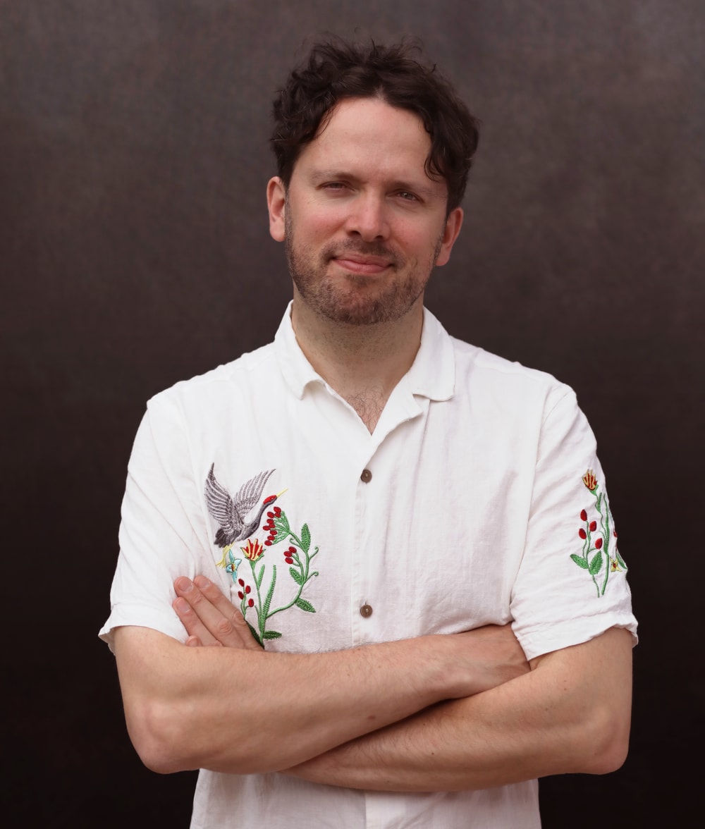 Mikey standing against a plain background. His arms are folded and he's smiling into the camera. He's wearing a white, short sleeved shirt with some flowers and birds embroidered onto the left hand side and the right sleeve.