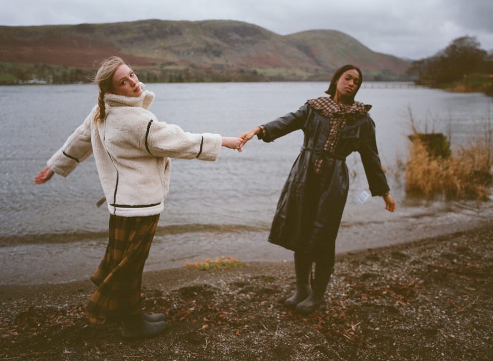 2 women standing leaning away from each other holding hands by a lake