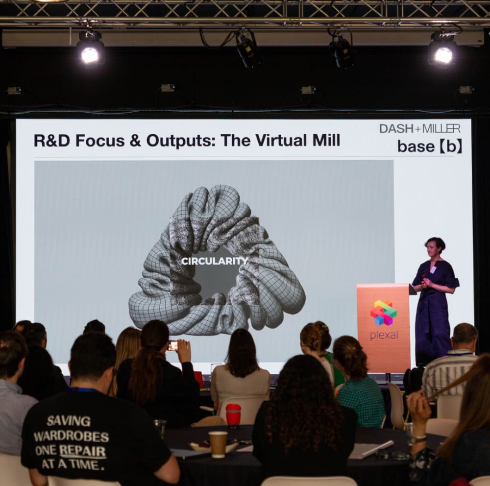 Steph Rolph, a woman with short hair standing on a stage with a large screen behind her presenting to an audience about research and development.
