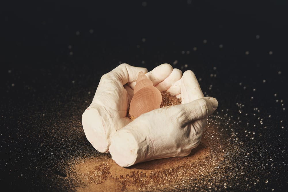 Plaster cast of a pair of hands holding a pink opaque shell and sand.