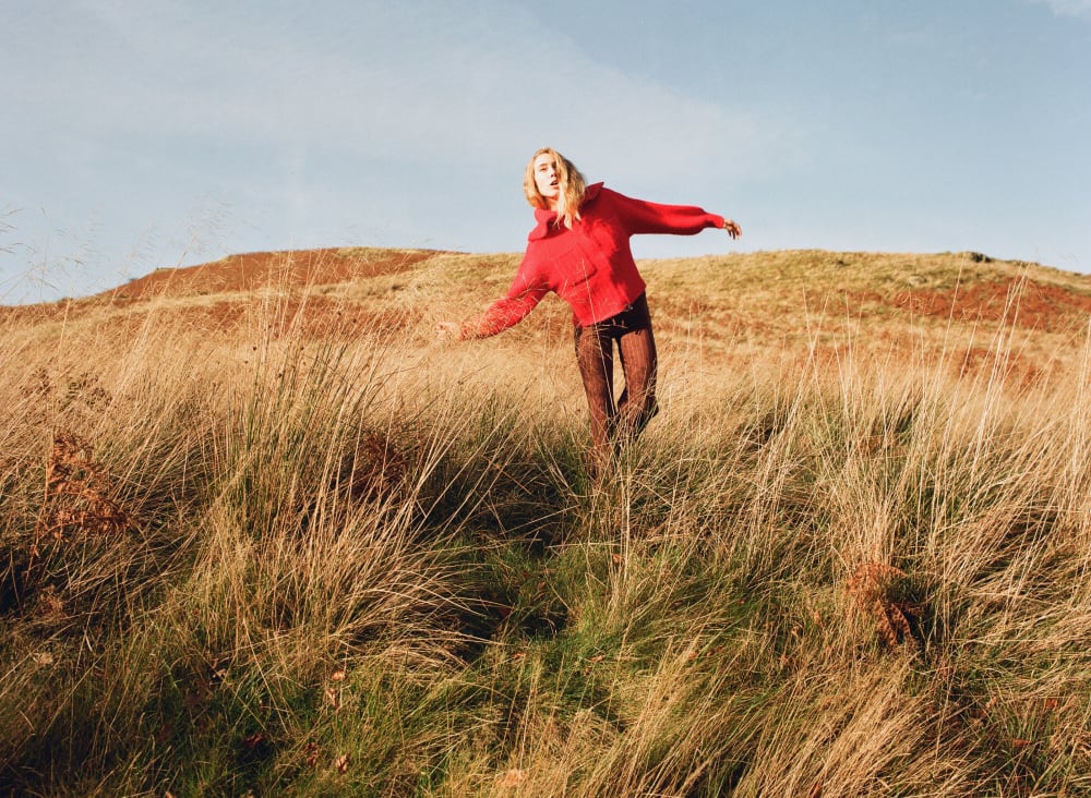 Woman posing in a field amidst dried long grass with hills in the background 